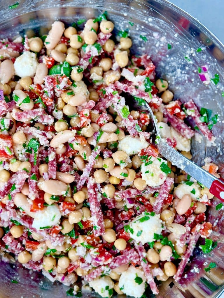 Close-up of La Scala dense bean salad recipe showing chickpeas, white beans, salami, mozzarella, parmesan, and fresh herbs coated in tangy vinaigrette.