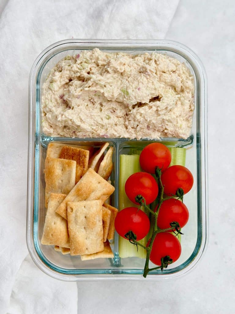 Round Swamp Farm chicken salad served with crackers, celery sticks, and cherry tomatoes in a glass meal prep container.