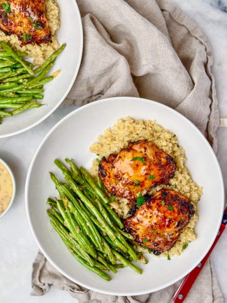 Two bowls of air fryer honey mustard chicken thighs with quinoa and green beans and dipping sauce on the side