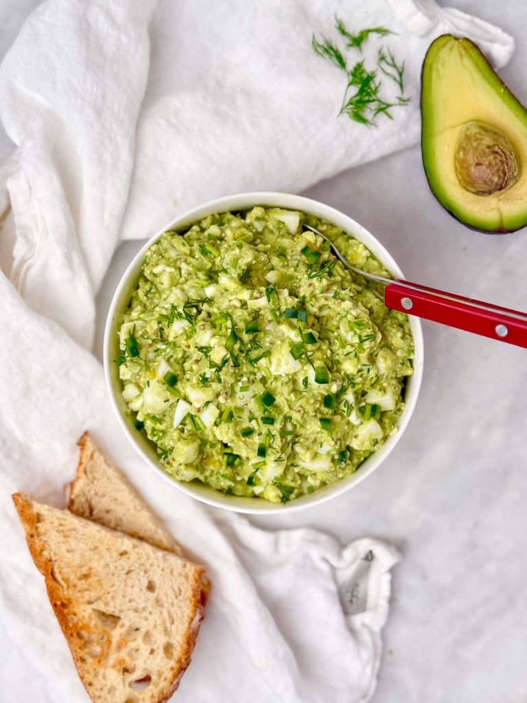 avocado egg salad in a white bowl overhead with toasted bread and halved avocado alongside
