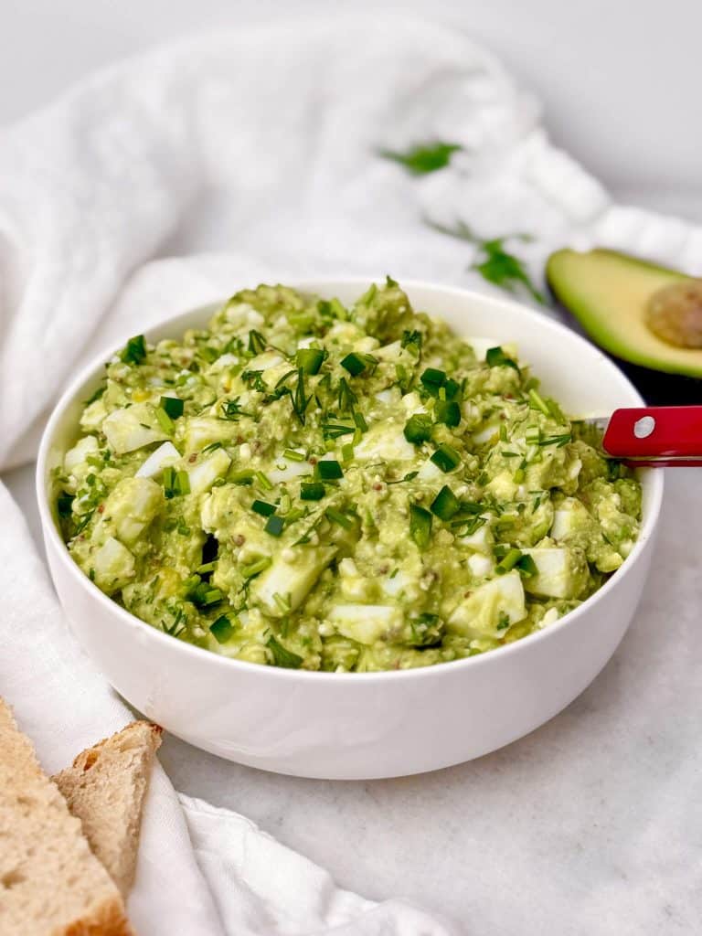 avocado egg salad in a white bowl topped with fresh chives and dill with toast and halved avocado alongside