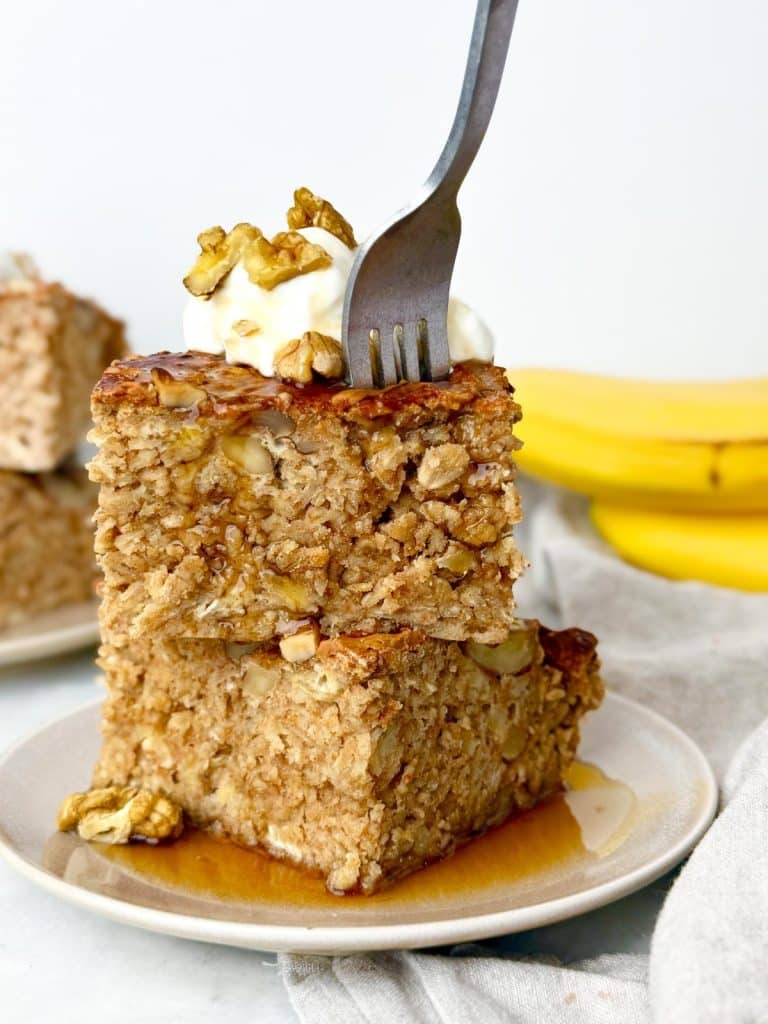 Fork pressing into a banana bread baked oatmeal square topped with yogurt and walnuts
