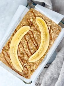 Unbaked banana bread oatmeal in a dish topped with banana halves and peanut butter drizzle