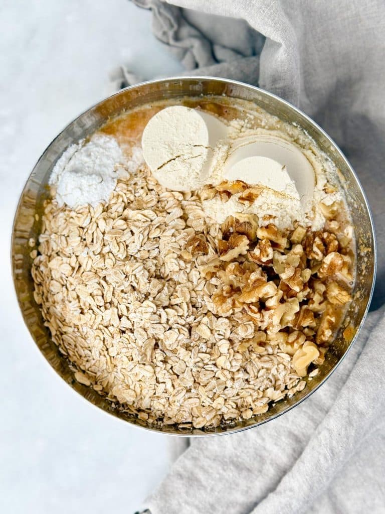 Rolled oats, walnuts, and protein powder added to banana batter in a mixing bowl