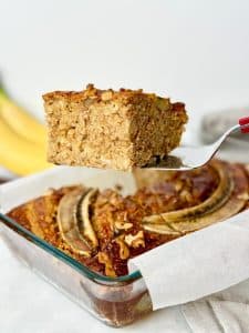 Slice of banana bread baked oats lifted from the baking dish with bananas in the background
