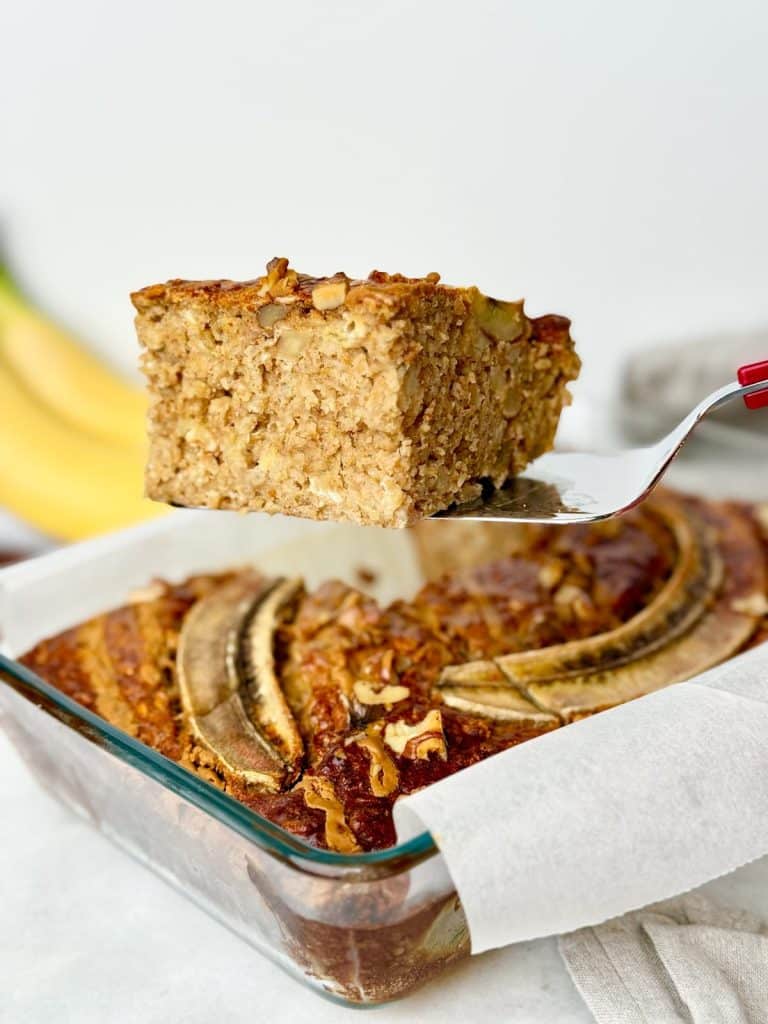 Slice of banana bread baked oats lifted from the baking dish with bananas in the background