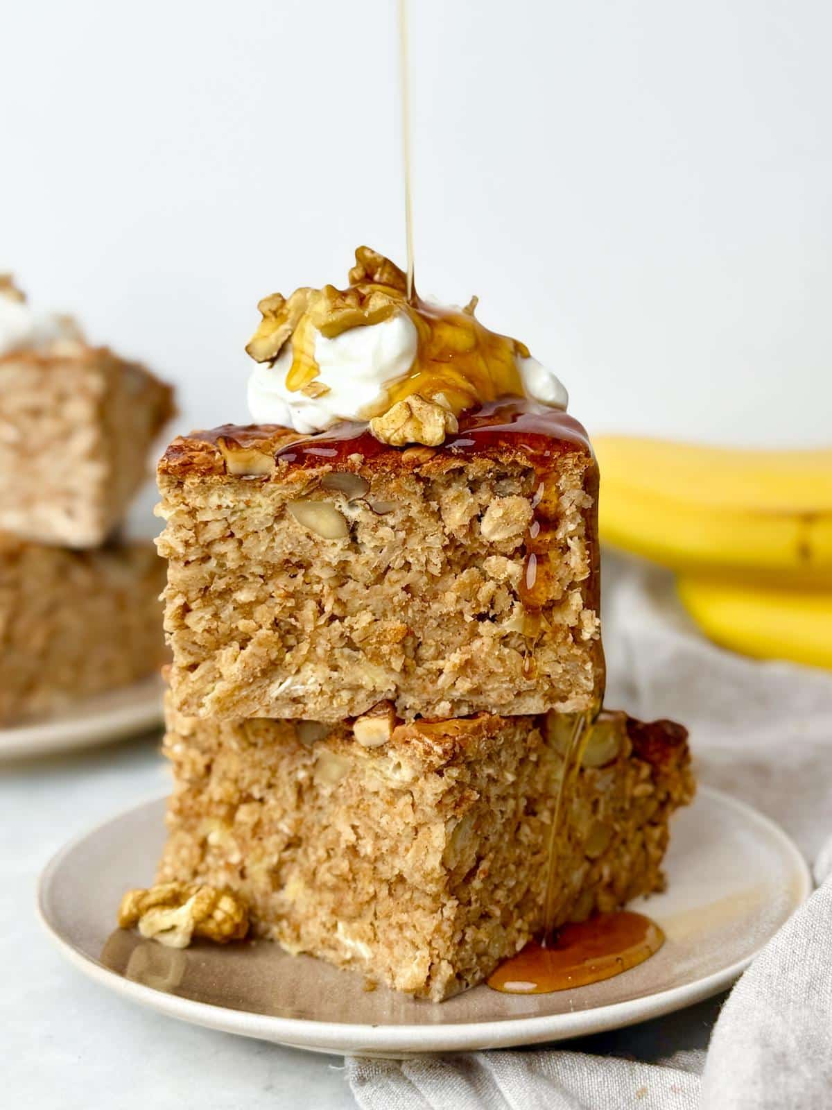 Banana bread baked oat squares stacked on a plate with maple syrup, yogurt, and walnuts