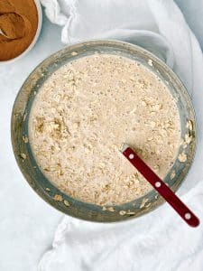 overnight oats batter with rolled oats stirred into the banana yogurt protein mixture in a large metal mixing bowl with a red spoon and cinnamon in background