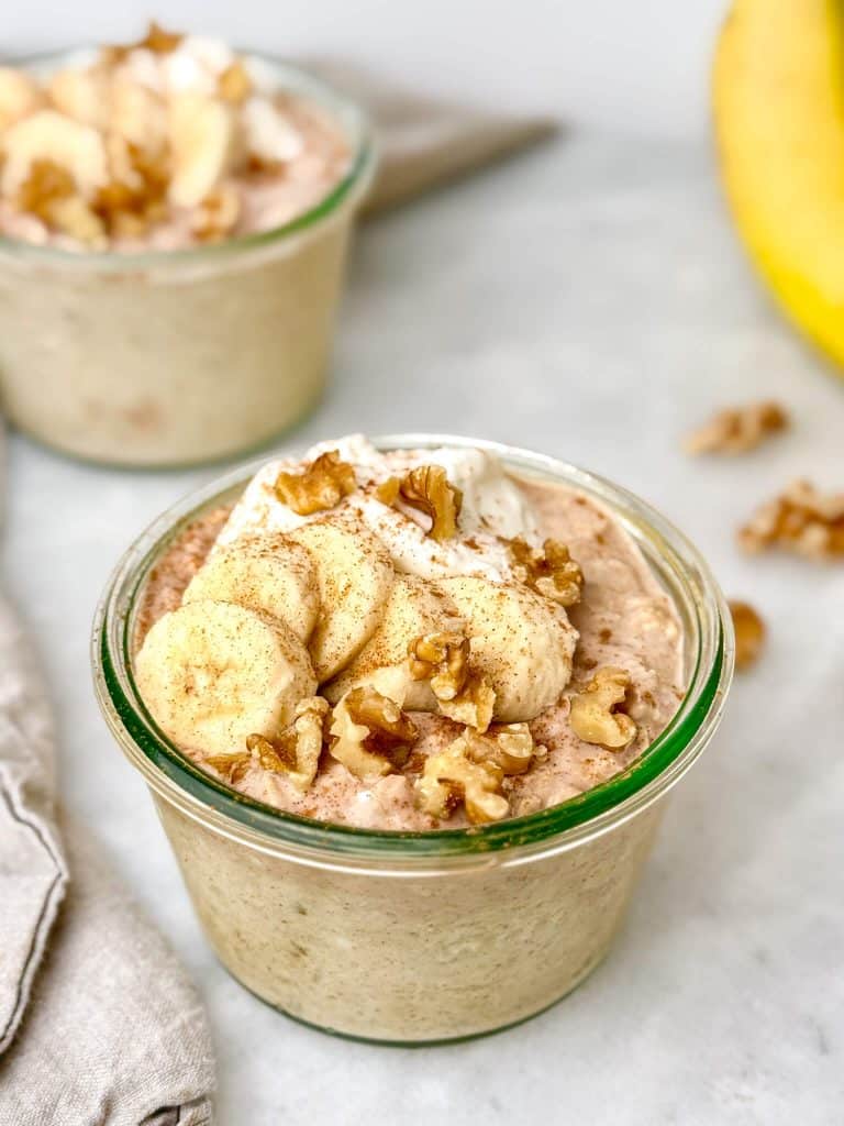 close up of banana bread overnight oats in a glass weck jar topped with banana slices, walnuts, Greek yogurt, and a dusting of cinnamon on a marble surface