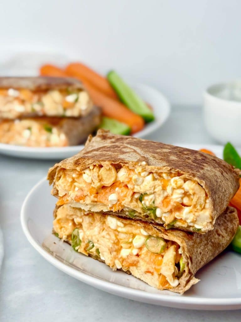 buffalo chicken wrap cut in half and stacked on a white plate with carrot and cucumbers.