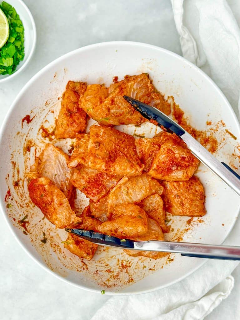 Cubed white fish coated in chili-lime seasoning with tongs in a white bowl