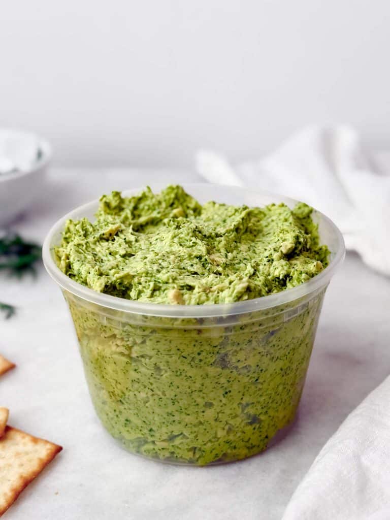 Green goddess chicken salad in a deli container with crackers and fresh herbs.