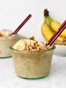 high protein banana bread overnight oats in a glass jar with a red spoon topped with banana slices and chopped walnuts with fresh bananas in the background