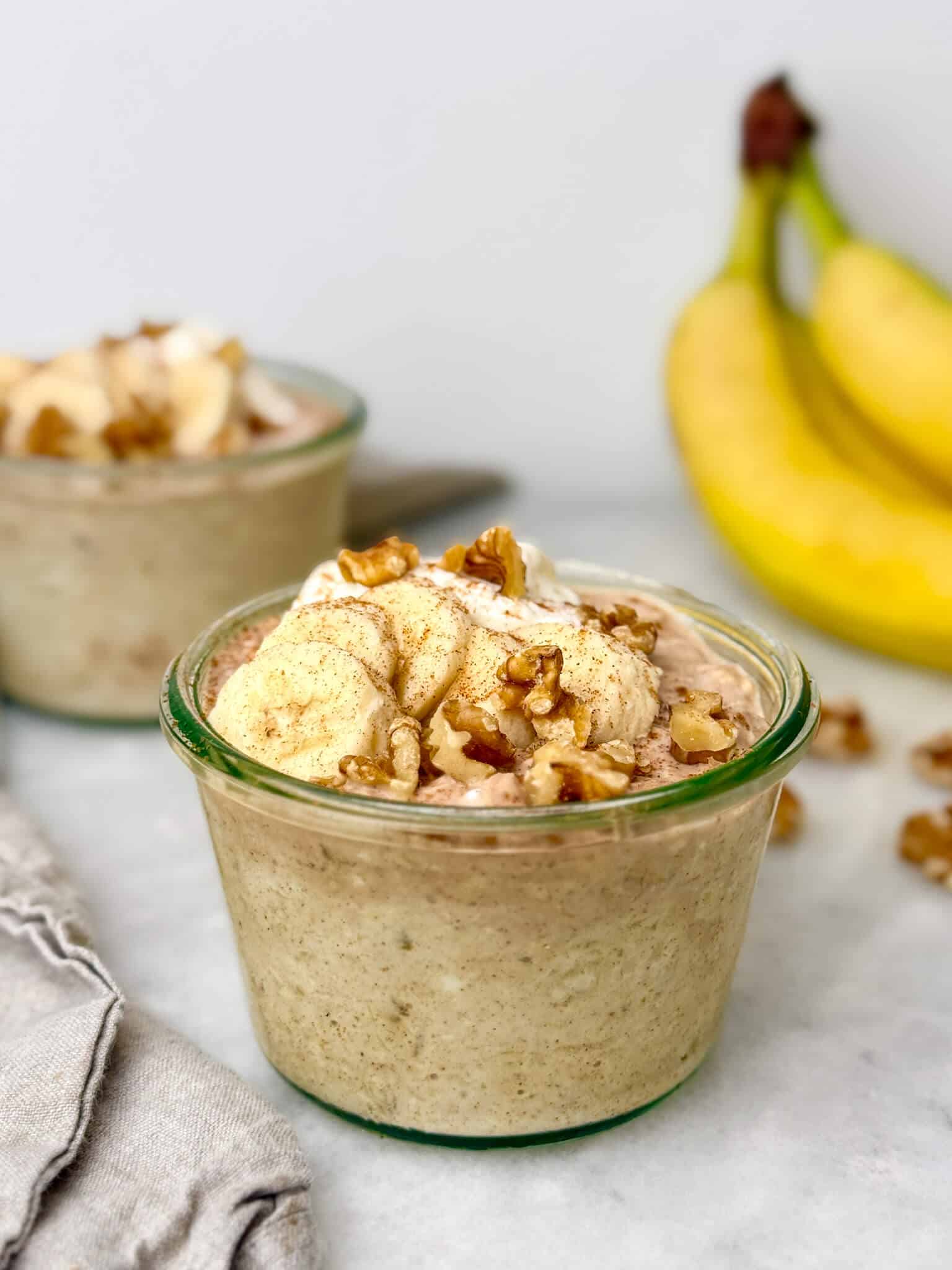 high protein banana bread overnight oats in a glass weck jar topped with banana slices, chopped walnuts, Greek yogurt, and cinnamon with a second jar and fresh bananas in the background