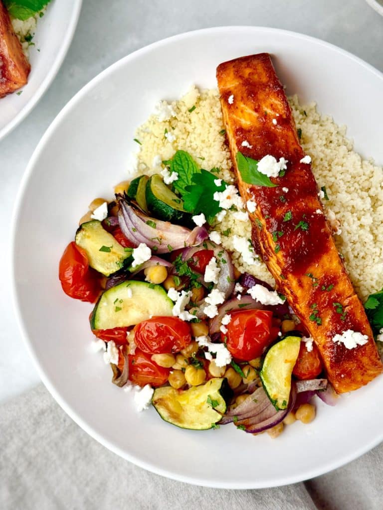 High protein honey harissa salmon bowl with couscous, roasted vegetables, chickpeas, fresh mint, parsley, and feta