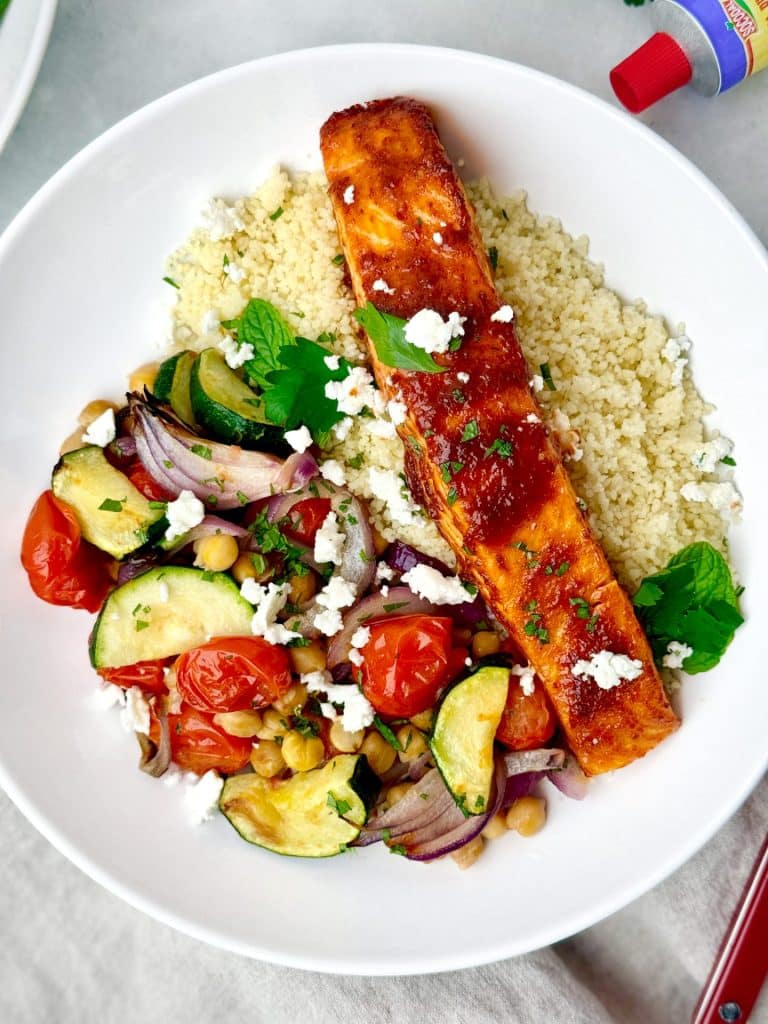 Close up of honey harissa air fryer salmon fillet over roasted zucchini, cherry tomatoes, chickpeas, and crumbled feta with couscous