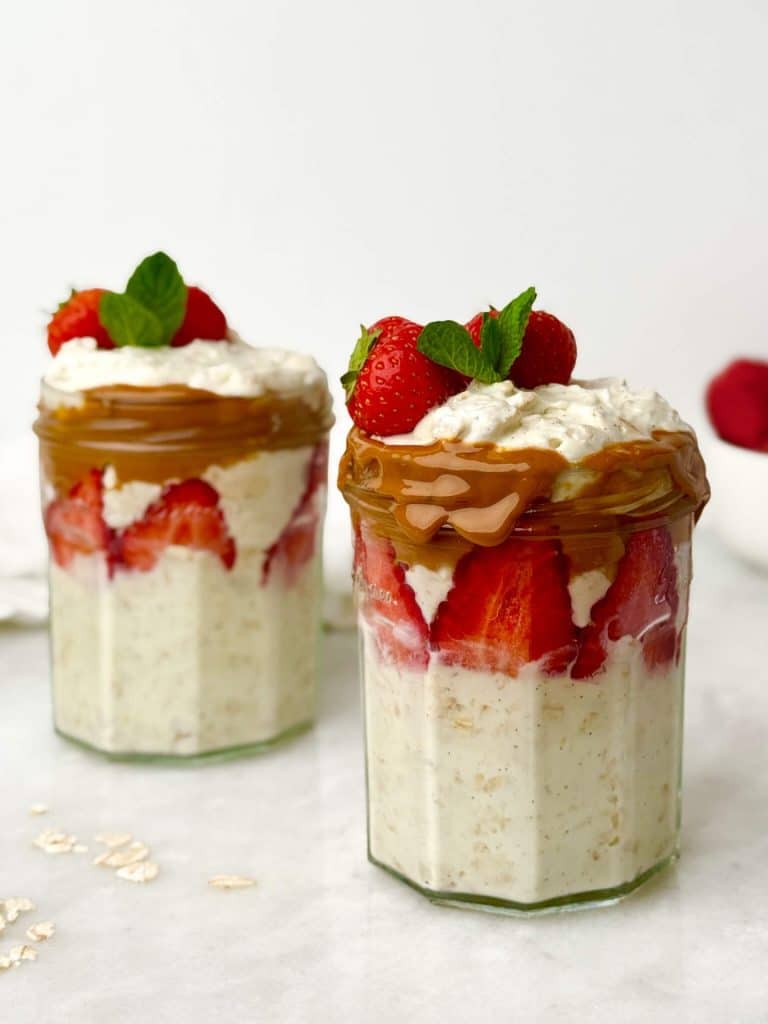 Two high-protein strawberry oat yogurt parfaits in individual glass jars with peanut butter drizzle and fresh strawberries, ready for breakfast meal prep.