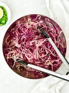 Shredded red cabbage slaw with tongs in a stainless steel bowl
