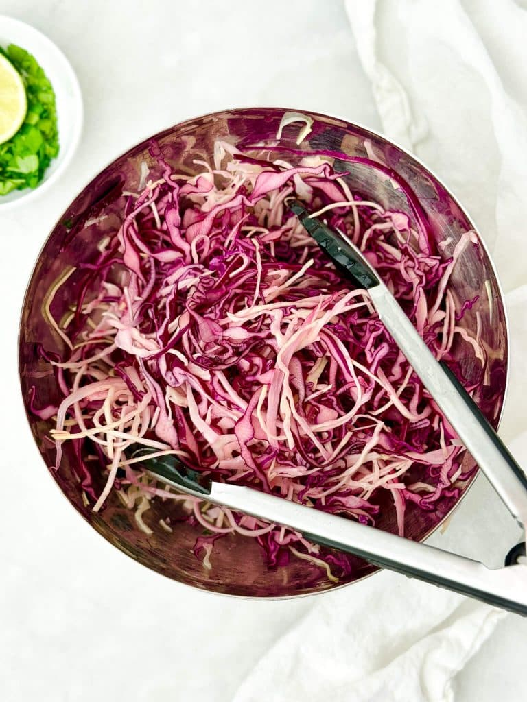 Shredded red cabbage slaw with tongs in a stainless steel bowl