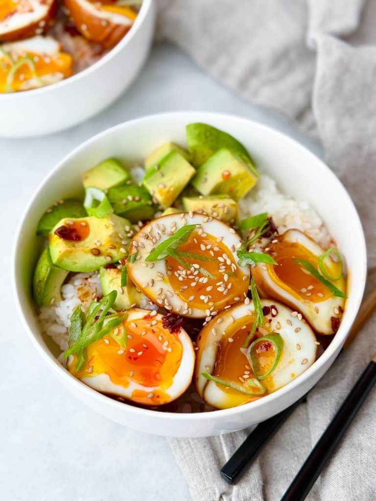 Jammy soy eggs over sushi rice with diced avocado, sesame seeds, and wooden chopsticks