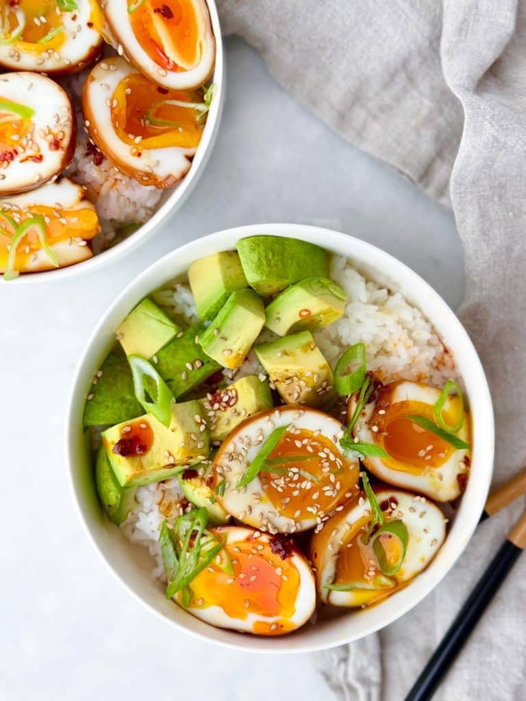 Two soy sauce egg rice bowls with halved jammy eggs, avocado, and chopsticks
