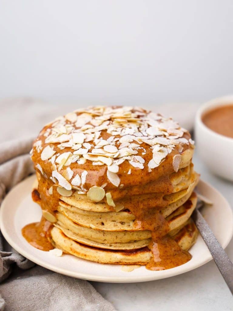 Full stack of almond croissant protein pancakes topped with almond cream drizzle and toasted sliced almonds on a white plate
