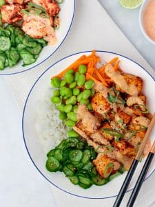 Two bang bang salmon bowls served over rice with edamame and cucumbers