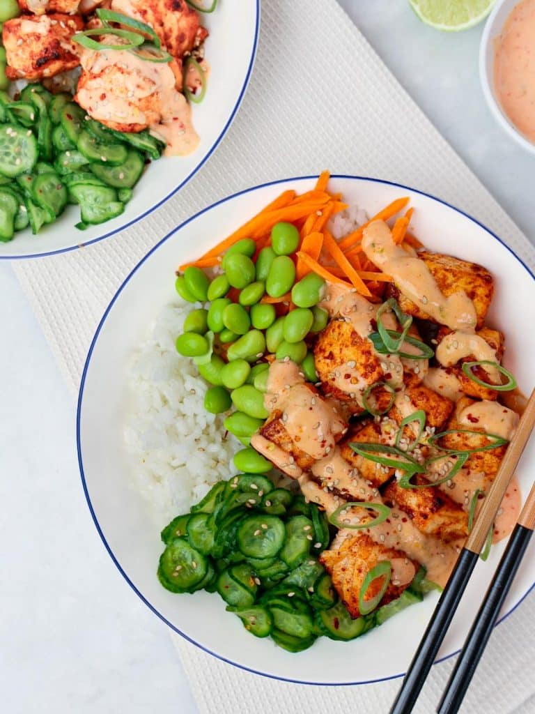 Two bang bang salmon bowls served over rice with edamame and cucumbers