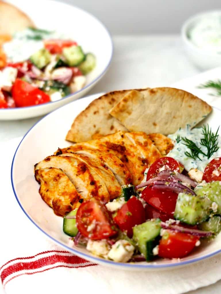 Angled view of chicken with tzatziki sauce, greek salad, and pita bread