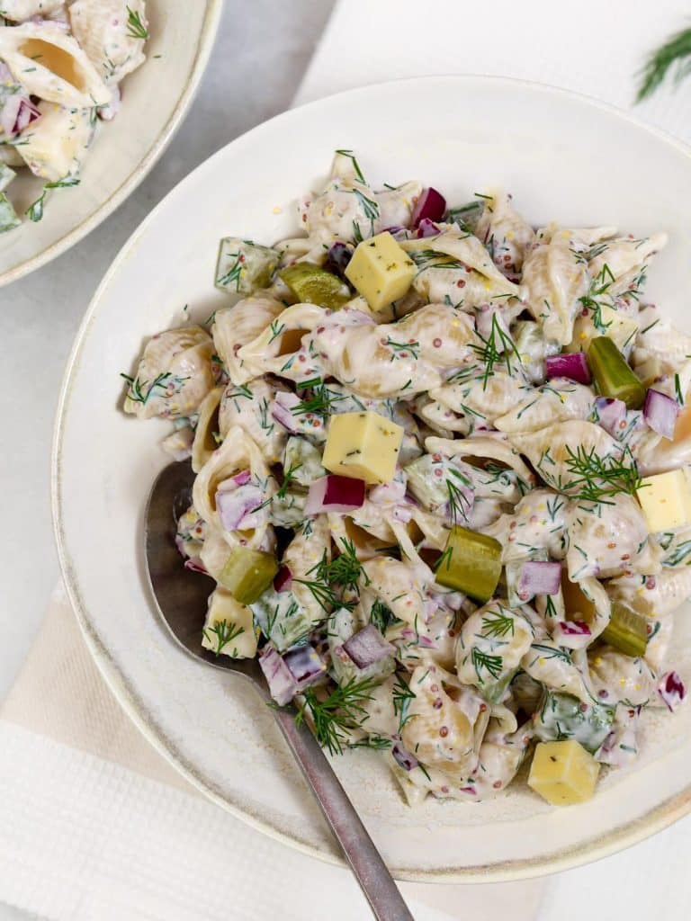 Healthy dill pickle pasta salad in a bowl with a spoon, loaded with red onion and havarti
