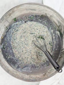 Creamy greek yogurt dressing being whisked with fresh dill and whole-grain mustard in a white bowl