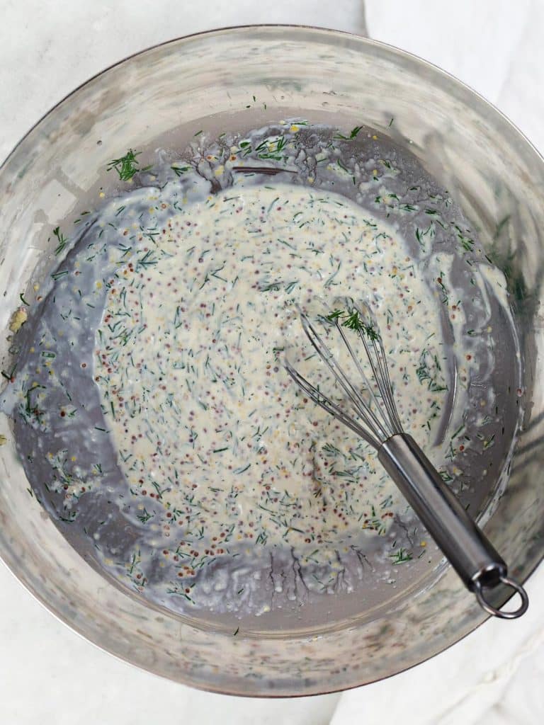 Creamy greek yogurt dressing being whisked with fresh dill and whole-grain mustard in a white bowl