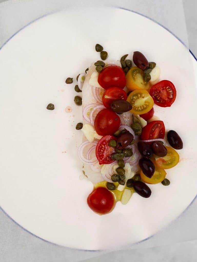 Raw cod fillet topped with shallot, garlic, cherry tomatoes, capers, and kalamata olives on parchment