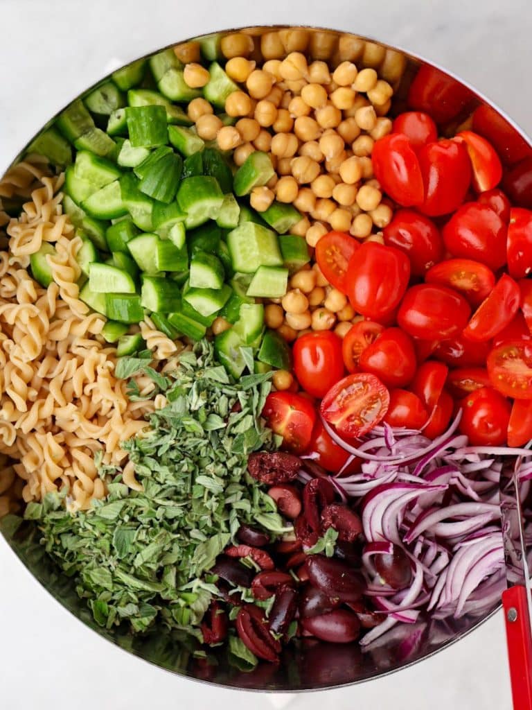 Mediterranean pasta salad ingredients including chickpeas, cherry tomatoes, cucumber, kalamata olives, red onion, and fresh oregano arranged in a bowl
