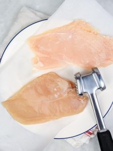 Two raw chicken breasts on a white plate next to a meat mallet, ready to be pounded thin for air fryer chicken katsu