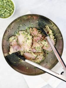 Raw chicken thighs tossed with fresh basil pesto in a stainless steel bowl