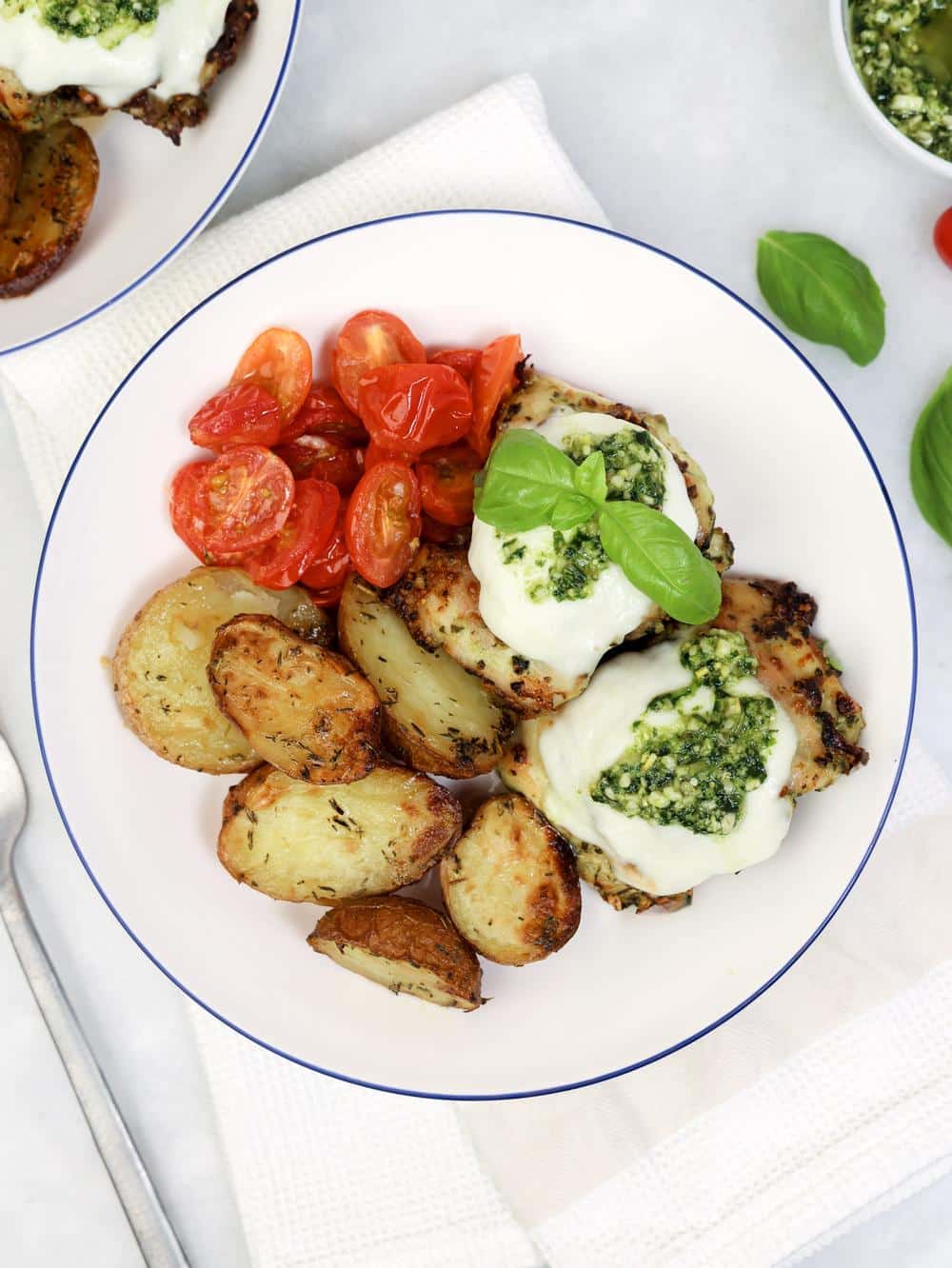 Plated air fryer pesto chicken with crispy garlic-rosemary potatoes, blistered cherry tomatoes, and fresh basil on a white plate