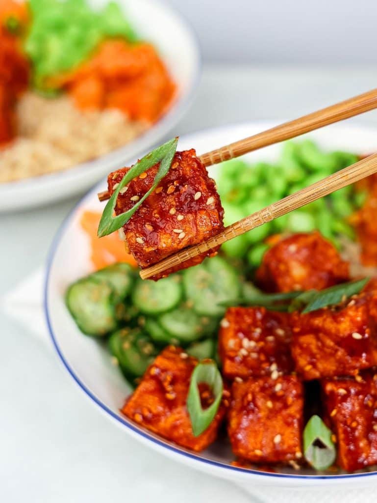 Close up of chopsticks holding a glazed gochujang tofu cube over a tofu bowl with cucumber