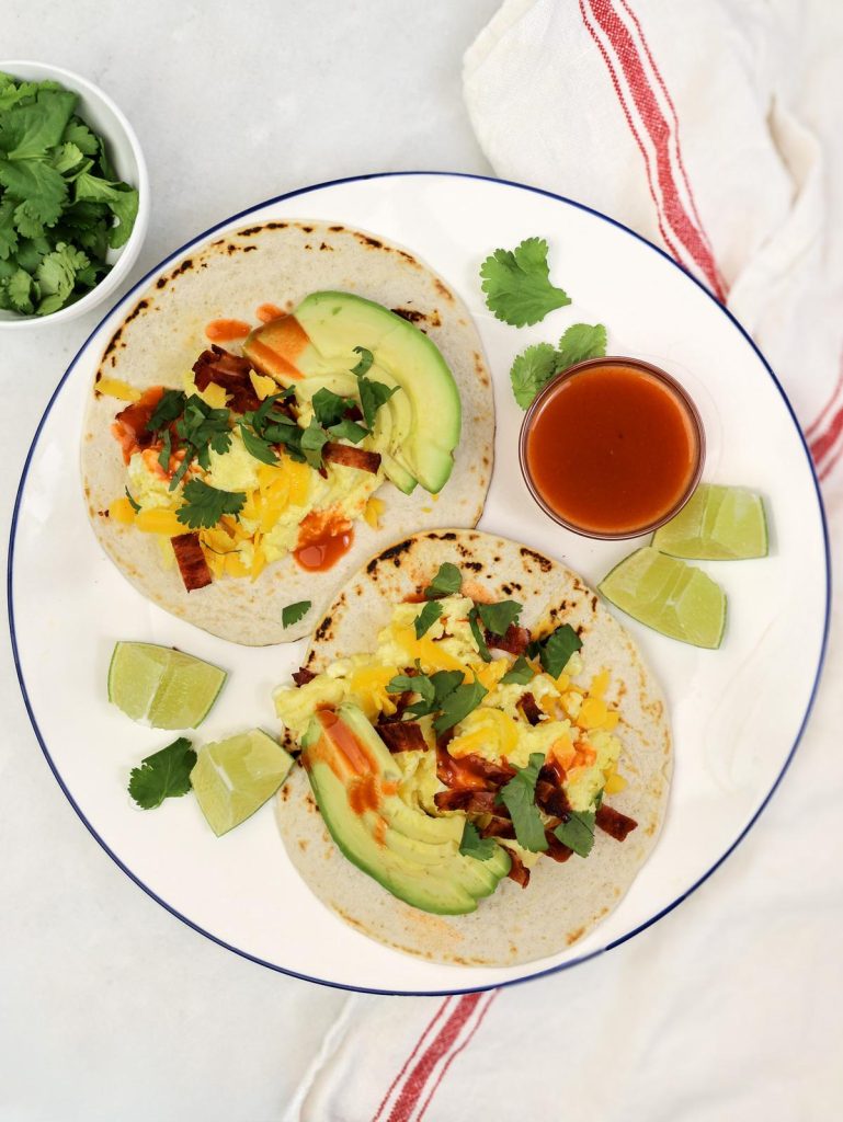 Two high-protein breakfast tacos on a plate with hot sauce, lime wedges, and fresh cilantro on the side