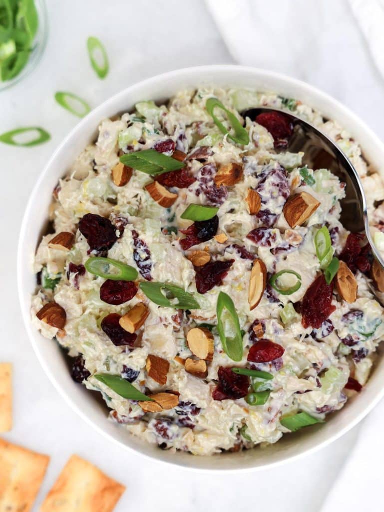 Chicken salad with dried cranberries and toasted almonds in a white bowl with a silver spoon, served with crackers