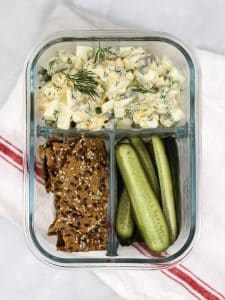 Spicy dill pickle egg salad packed in a glass meal prep container with seed crackers and pickle spears