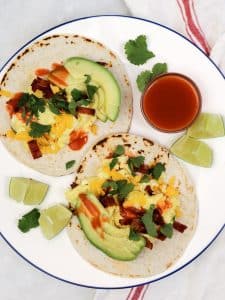 Two high-protein breakfast tacos with scrambled eggs, turkey bacon, cheddar, avocado, and hot sauce on a blue-rimmed plate