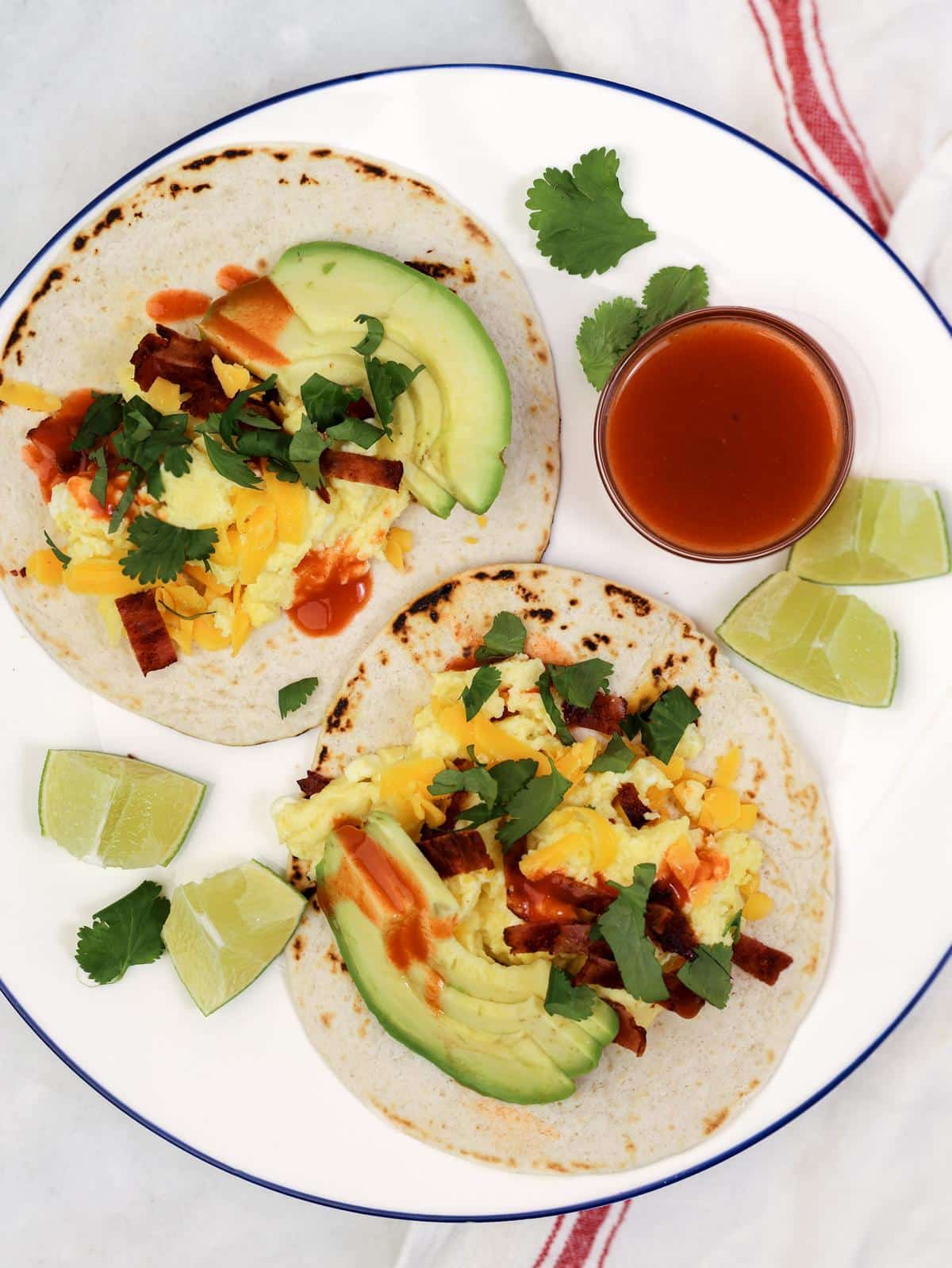 Two high-protein breakfast tacos with scrambled eggs, turkey bacon, cheddar, avocado, and hot sauce on a blue-rimmed plate