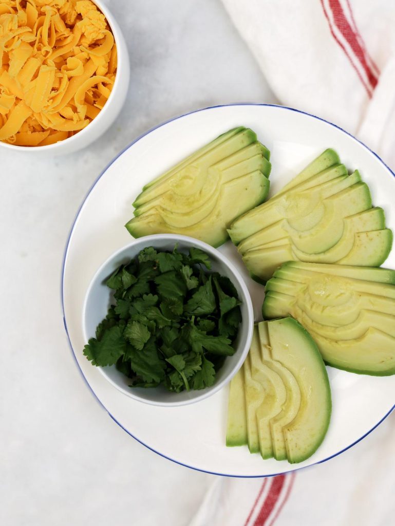 Sliced avocado, chopped fresh cilantro, and shredded sharp cheddar in small bowls, ready for assembling breakfast tacos