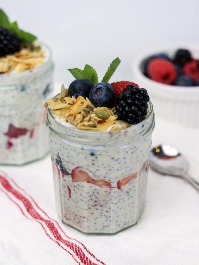 Overnight chia seed yogurt parfait in a glass jar with granola, mixed berries, mint, and a spoon on the side