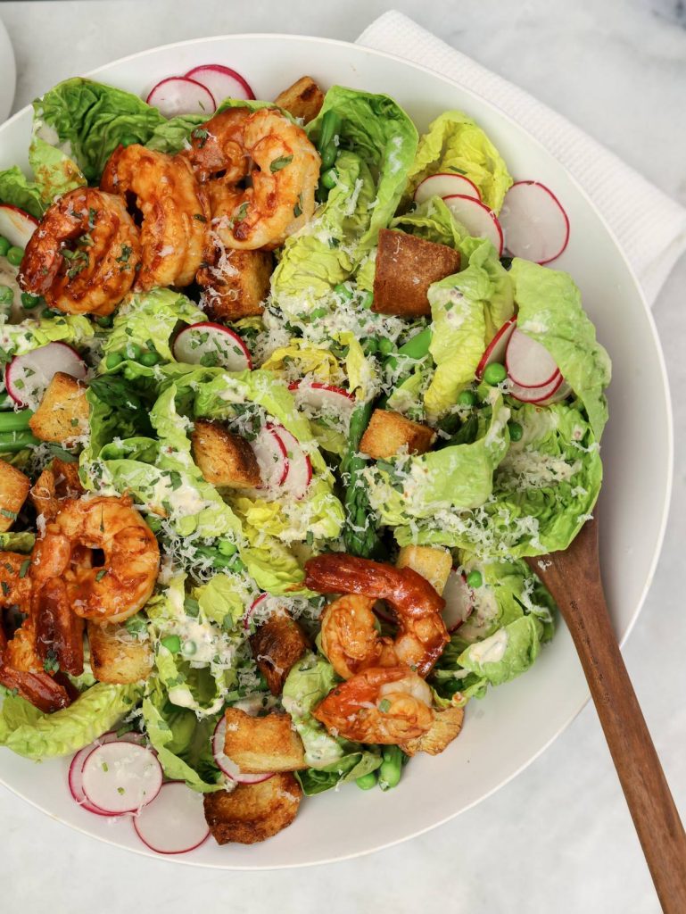 Spring caesar salad recipe served in a white bowl with shrimp, sliced radishes and a wooden serving spoon