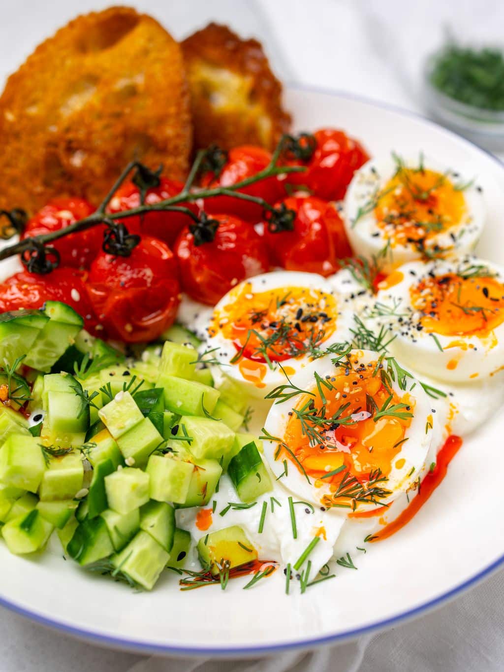 Close-up greek yogurt bowl with jammy eggs, hot honey drizzle, dill, and cucumber