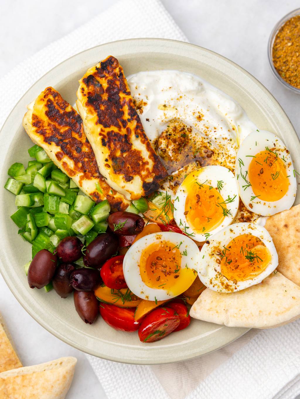 High-protein mediterranean breakfast plate with jammy eggs, halloumi, marinated tomatoes, olives, and za'atar yogurt