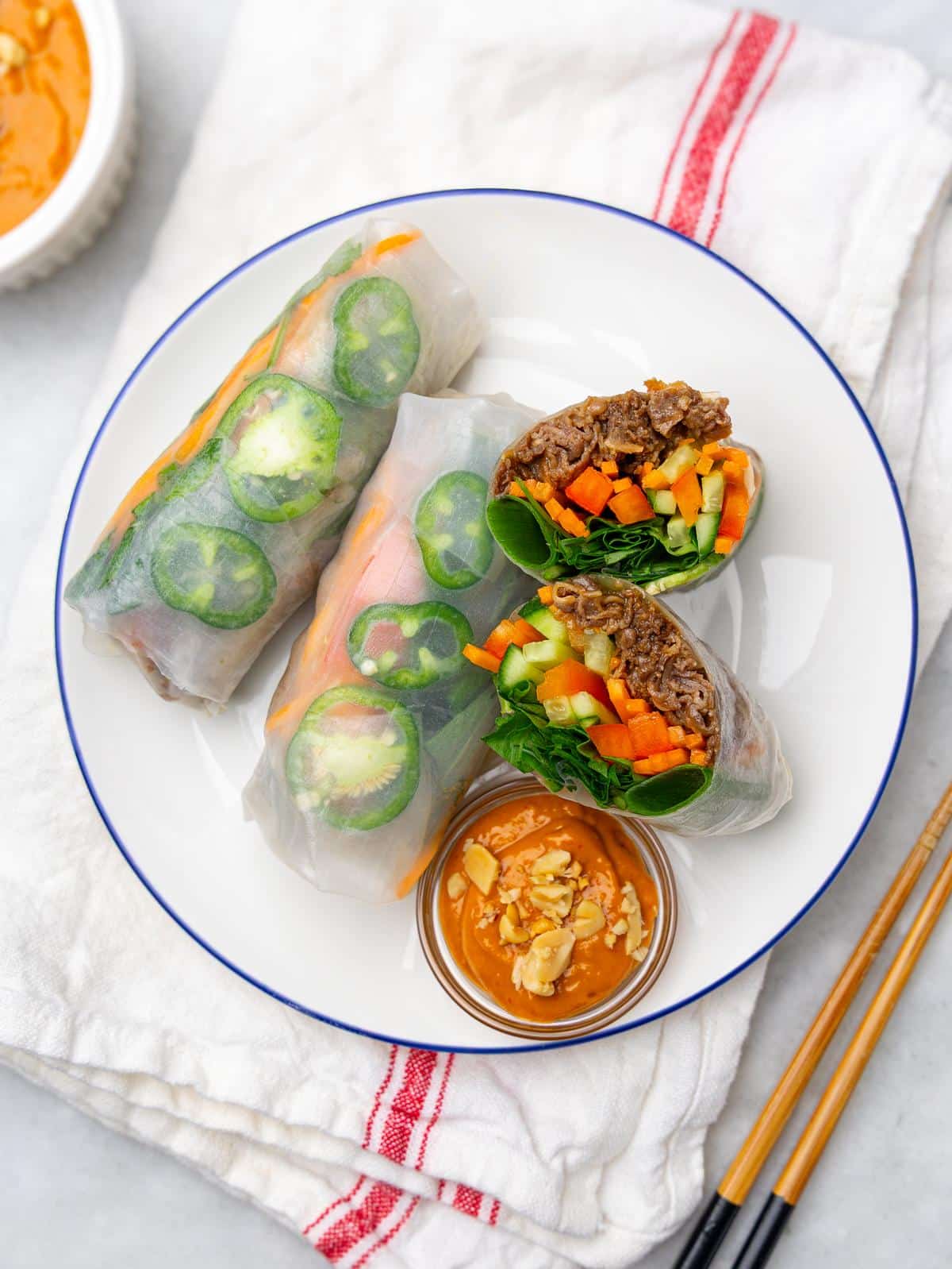 High-protein summer rolls on a plate with one cut open, jalapeño on top, and spicy peanut sauce