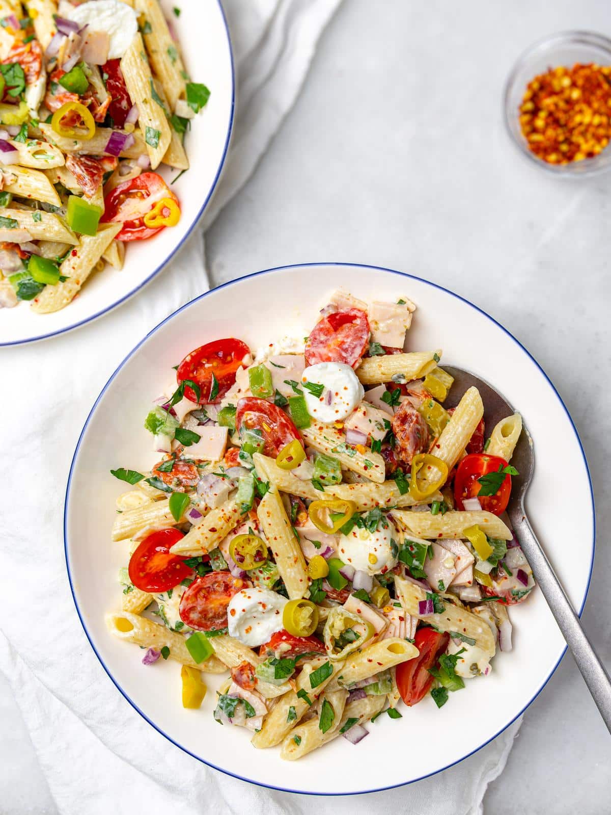 Two blue-rimmed bowls of italian grinder pasta salad with a spoon and red pepper flakes